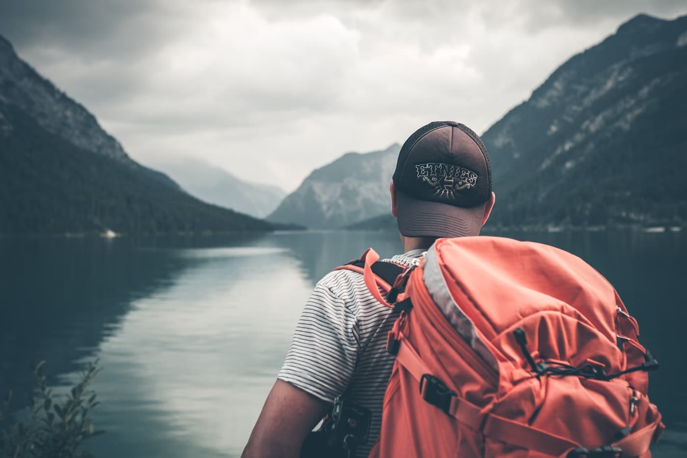 A hiker with an orange backpack facing a mountain lake